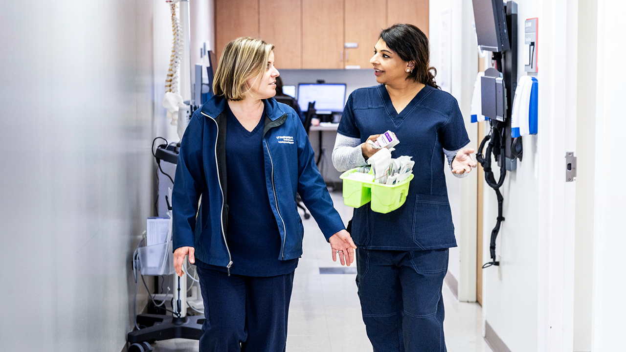 Two nurses walking down a hall talking to each other. One is holding a green container with equipment.
