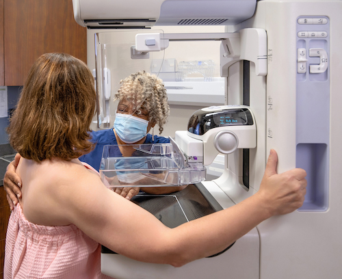 A healthcare professional wearing a mask assists a woman in a pink top during a mammogram.