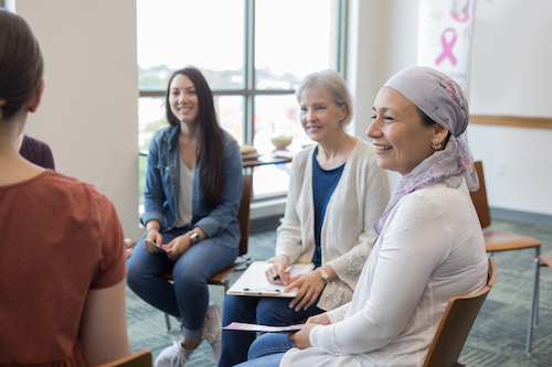 A group of diverse women, including one with a headscarf, sitting in a circle and smiling, representing a supportive and communal environment for cancer patients.