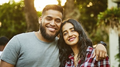 A man and woman standing close together, smiling at the camera, with greenery in the background.