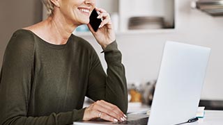 A smiling woman sitting at a table, talking on the phone while using a laptop.